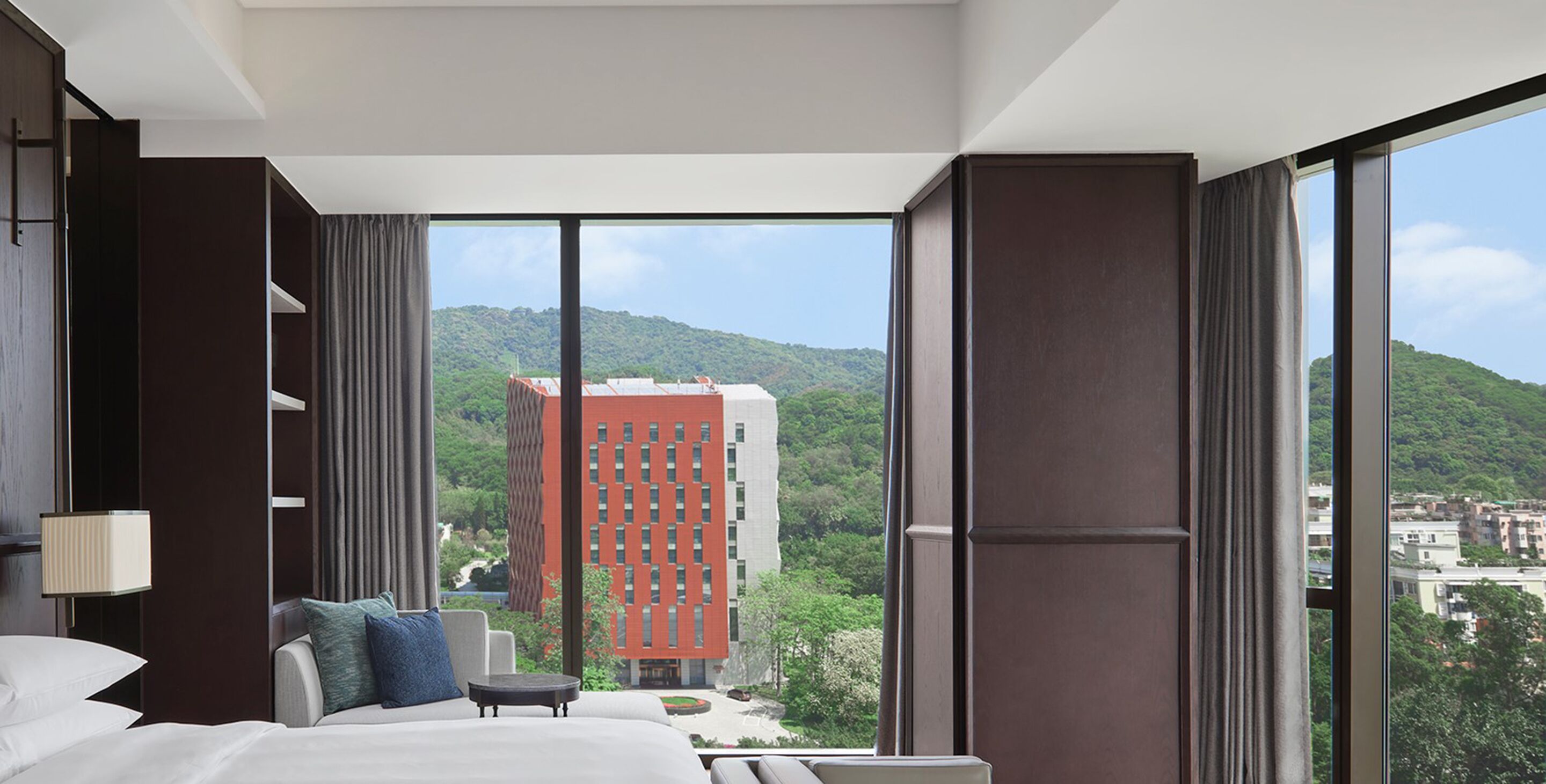 A modern hotel room with a large bed, a small table, and a view of a cityscape and mountains through floor-to-ceiling windows.