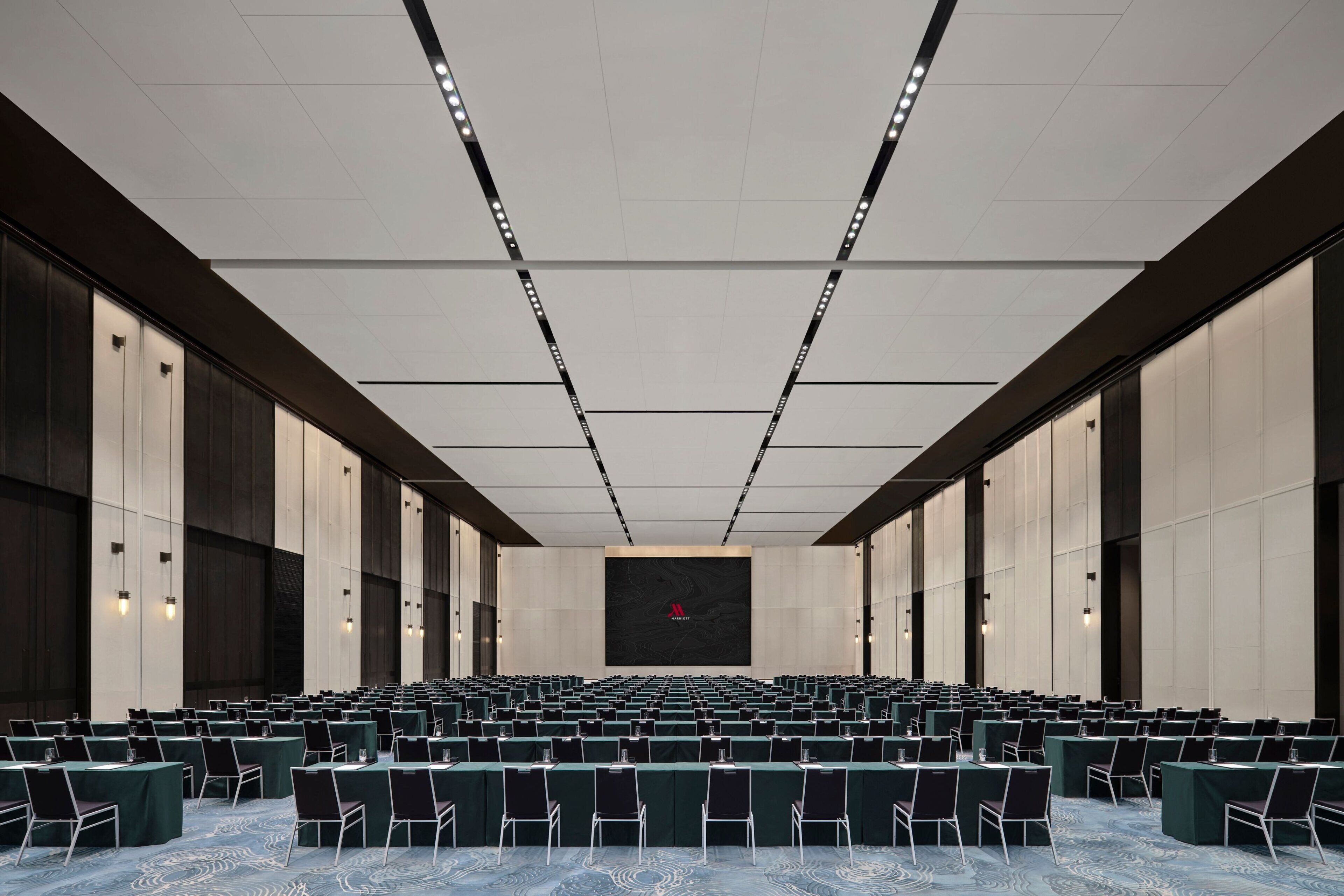 A conference room with rows of tables and chairs, a large screen at the front, and a modern ceiling design.
