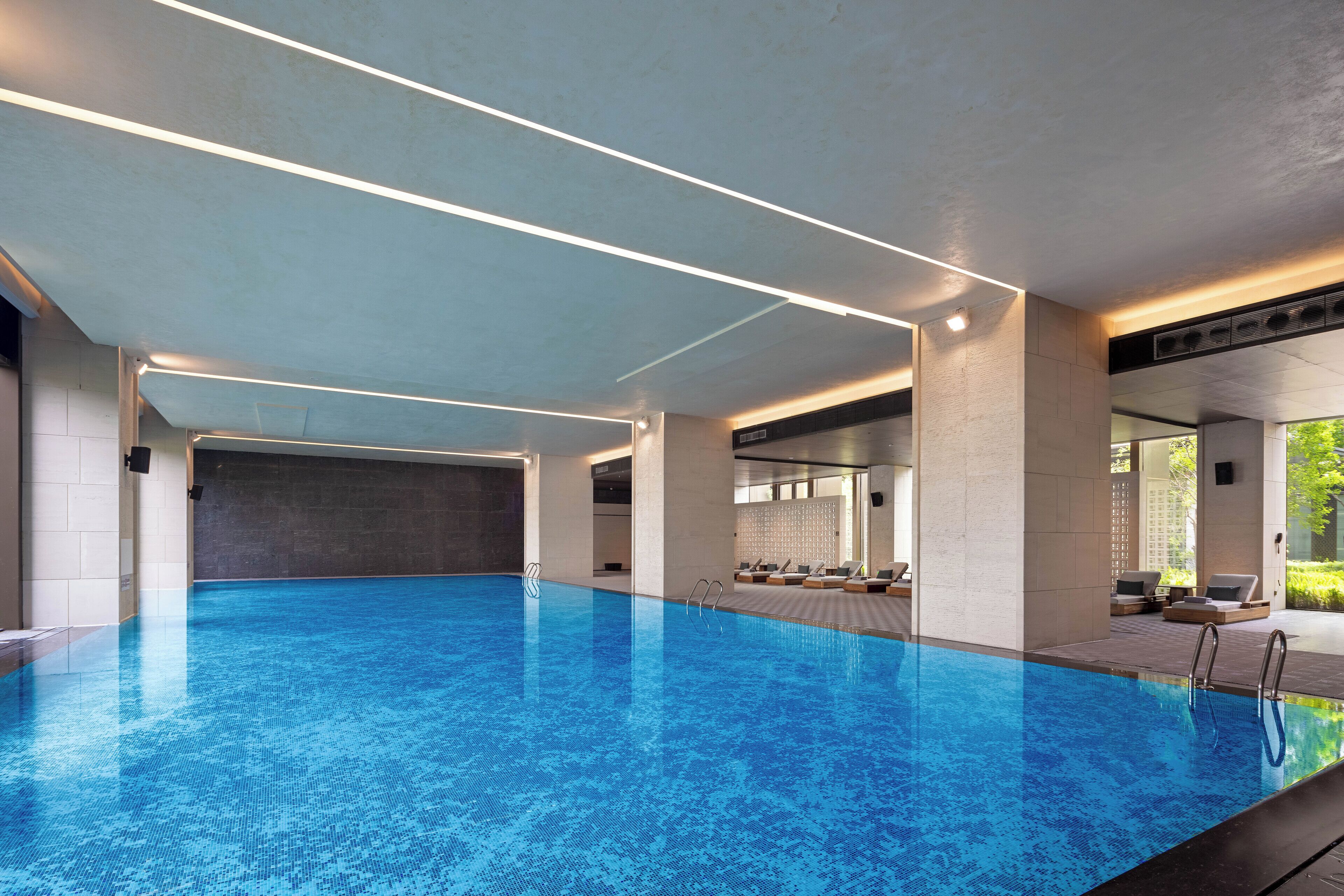 A large indoor swimming pool with clear blue water, surrounded by lounge areas with seating and a view of greenery outside.