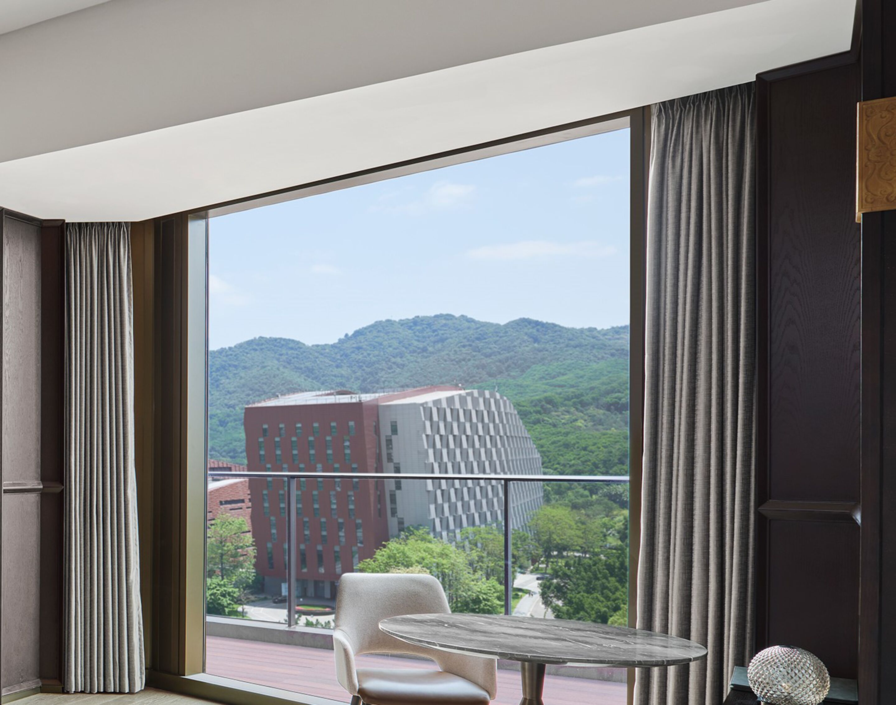 A modern hotel room with a large window offering a view of a cityscape and mountains.