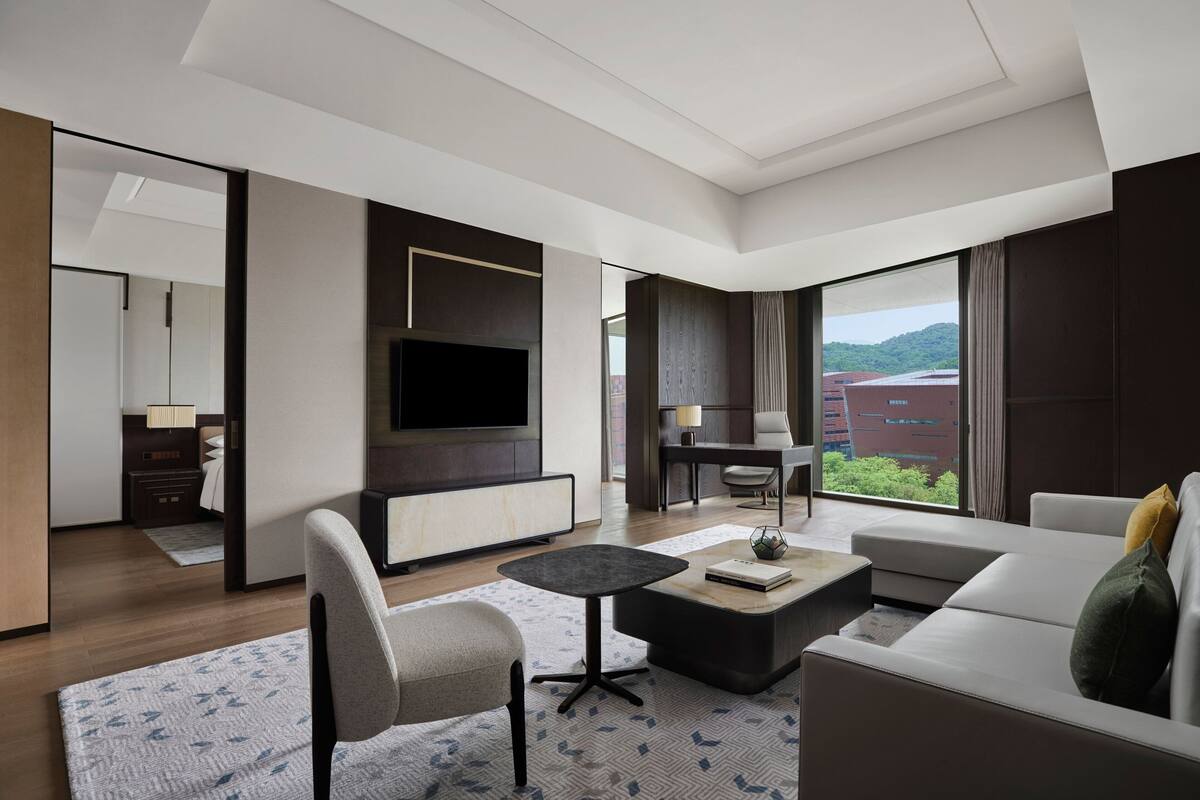 Une chambre d’hôtel moderne dotée d’une grande fenêtre donnant sur la ville, d’un téléviseur à écran plat fixé au mur, d’un canapé confortable, d’une petite table basse et d’un coin salon avec un fauteuil et une lampe.