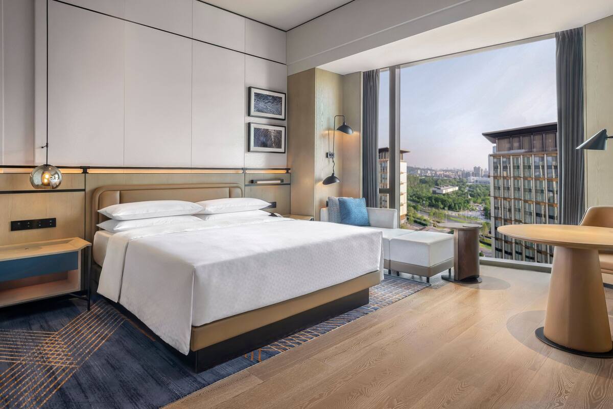 A modern hotel room with a large bed, a small table, a chair, and a view of the city through large windows.