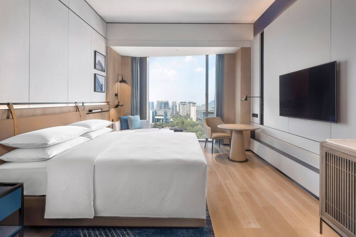 A modern hotel room with a large bed, a TV, a small table, and a view of the city.