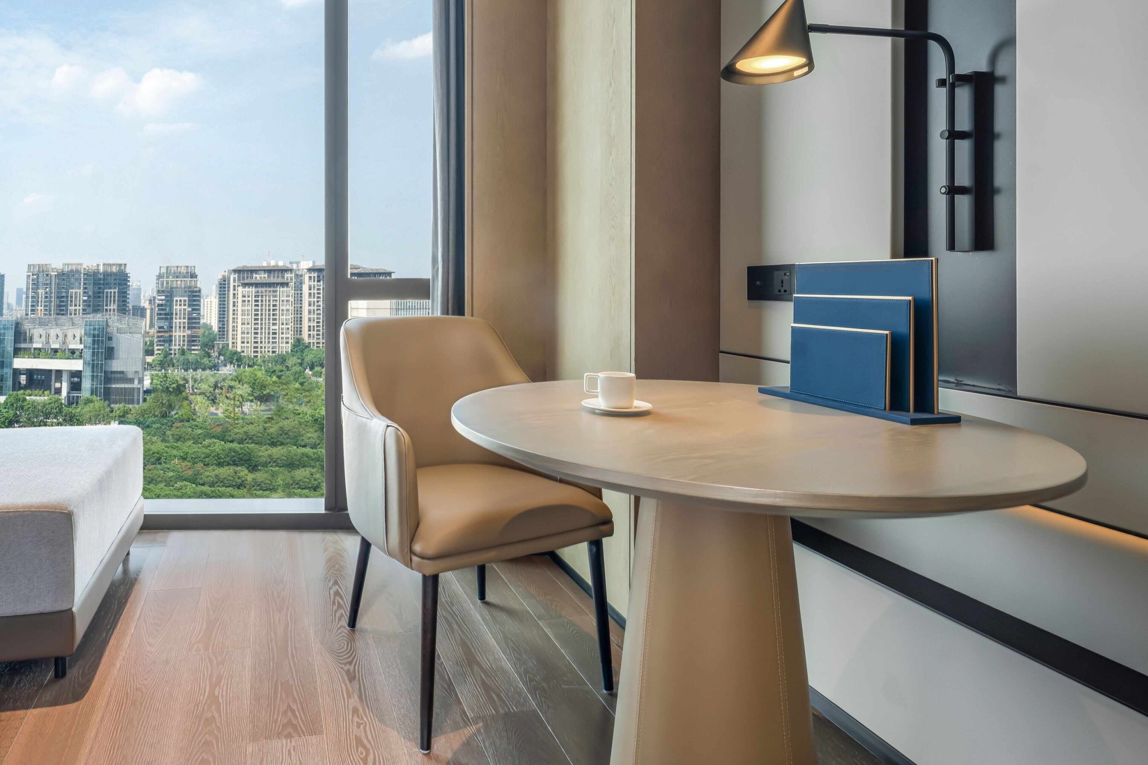 A modern hotel room with a round table, a chair, a bedside table, a lamp, and a city view through large windows.