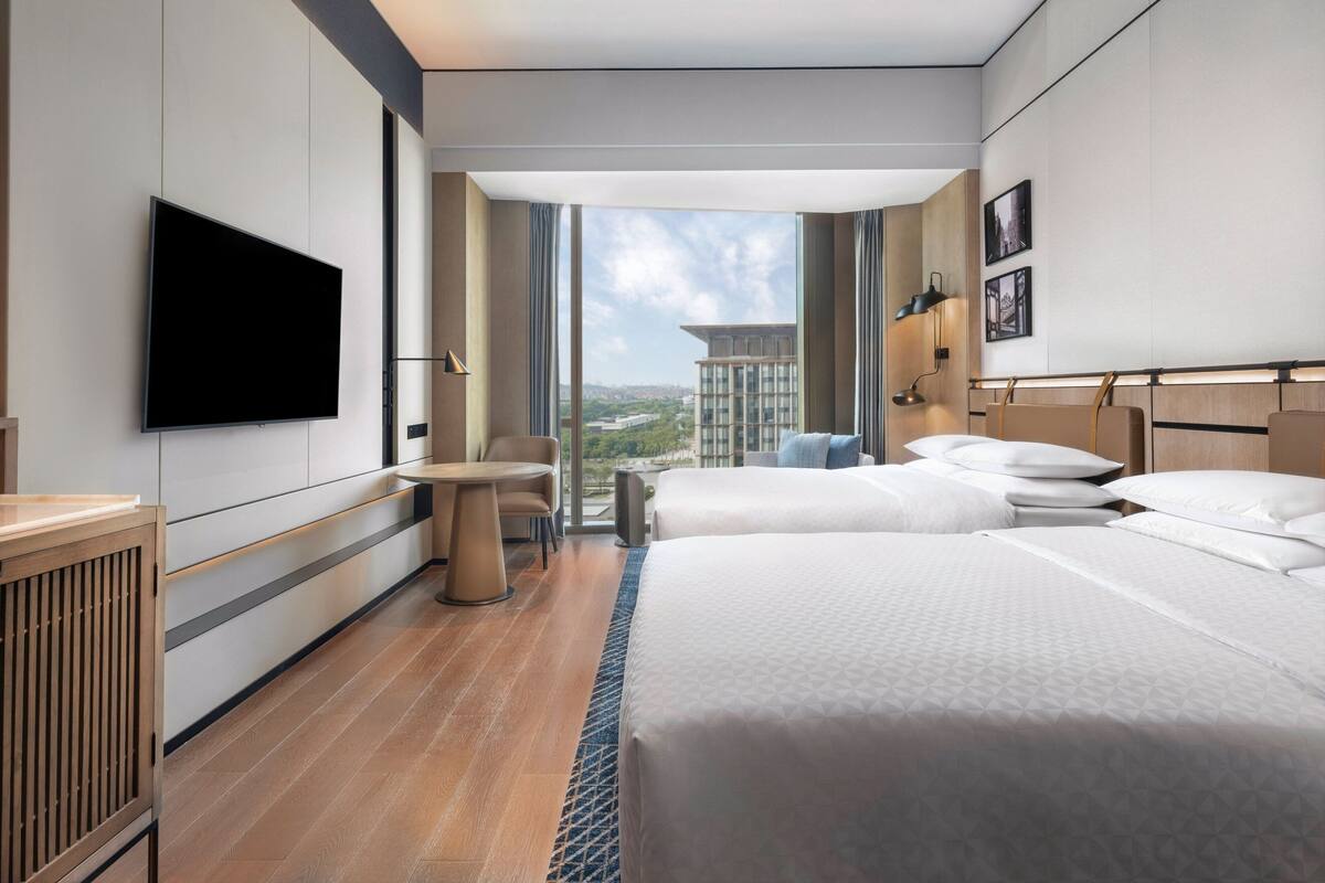 A modern hotel room with two beds, a flat-screen TV, a desk, and a large window offering a city view.