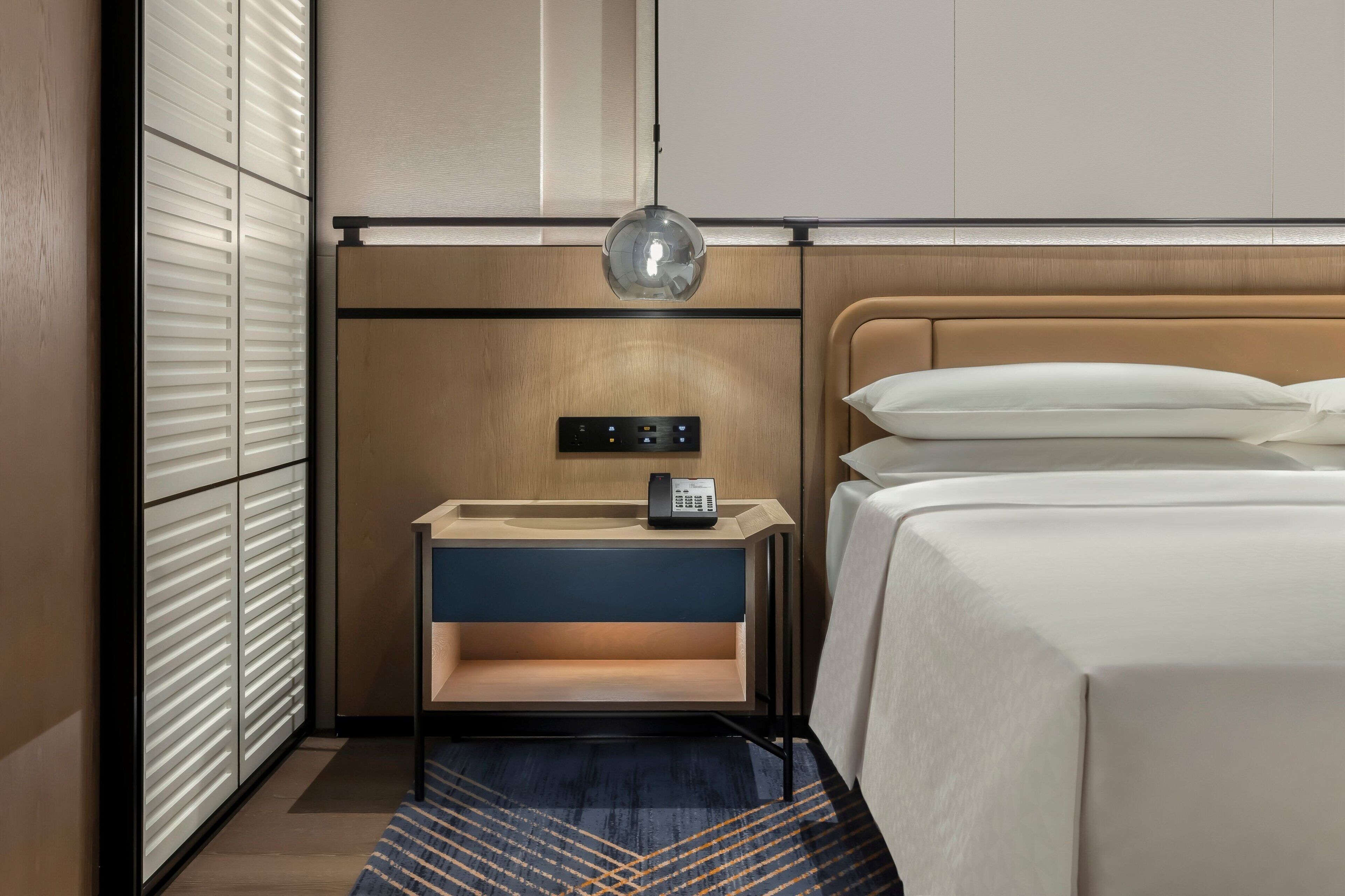 A hotel room with a bed, bedside table, lamp, and a control panel on the wall.