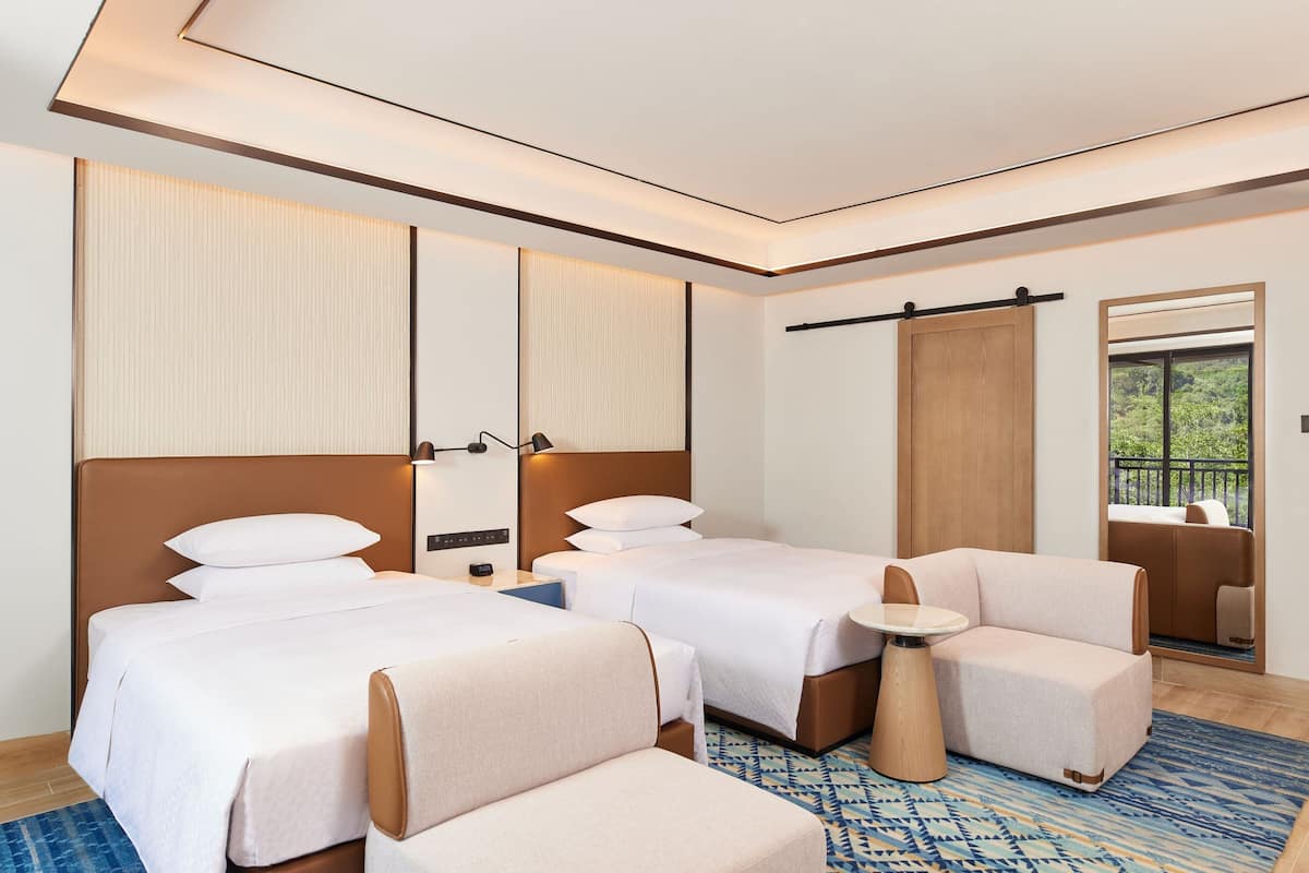 Habitación de hotel con dos camas, un sofá, una mesita y un ventanal con vistas a zonas verdes.