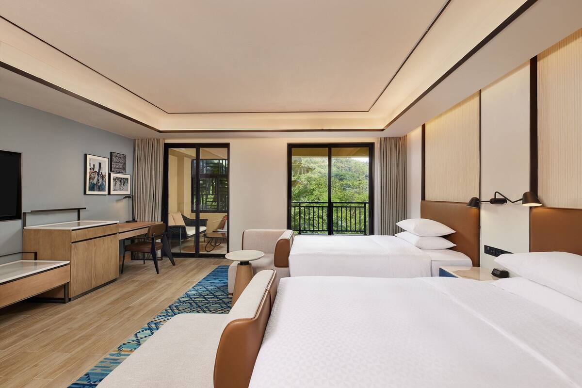 Habitación de hotel moderna con dos camas, un escritorio, una silla, un televisor y un balcón con vistas a zonas verdes.