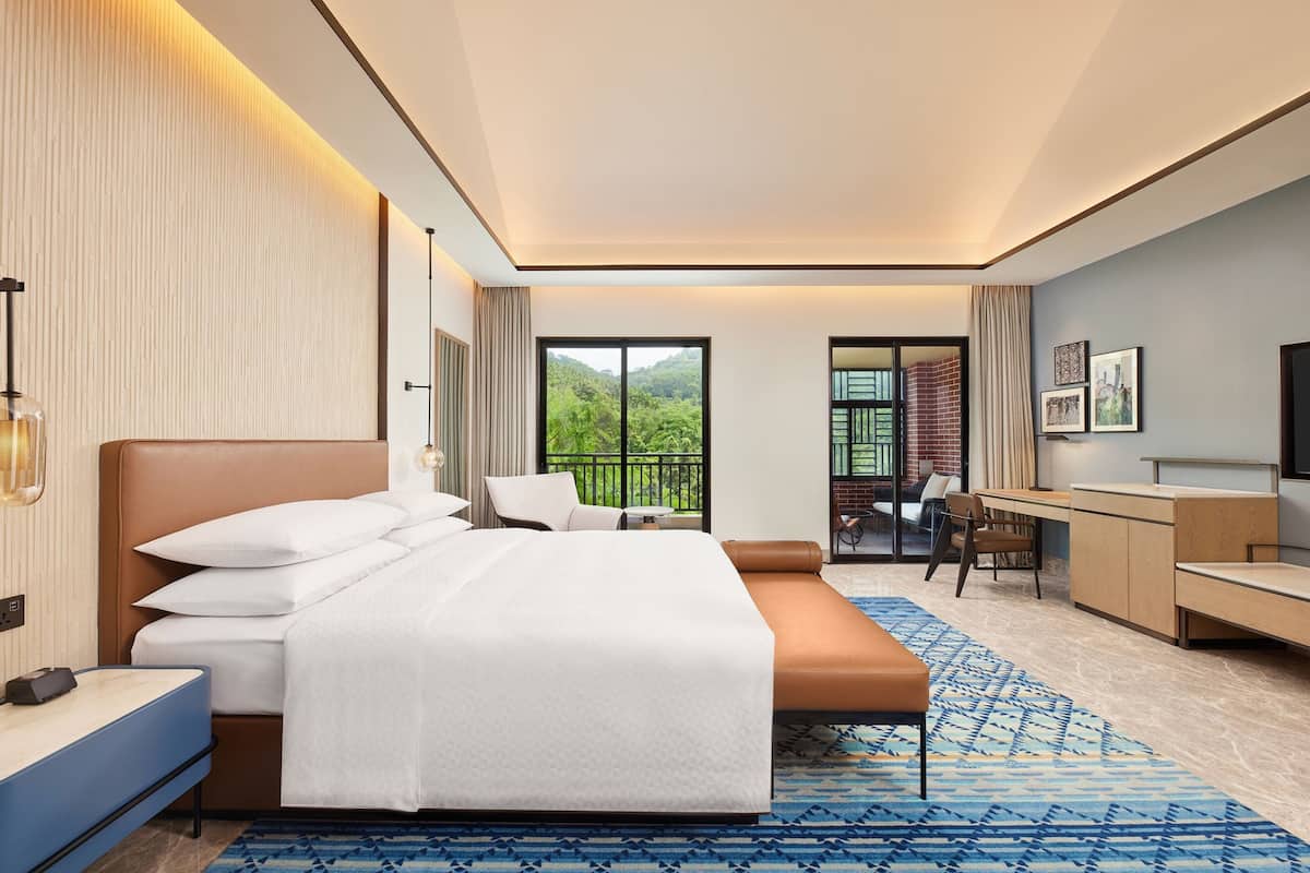 Una habitación de hotel moderna con una cama grande, un escritorio, una silla y un balcón con vistas a zonas verdes.