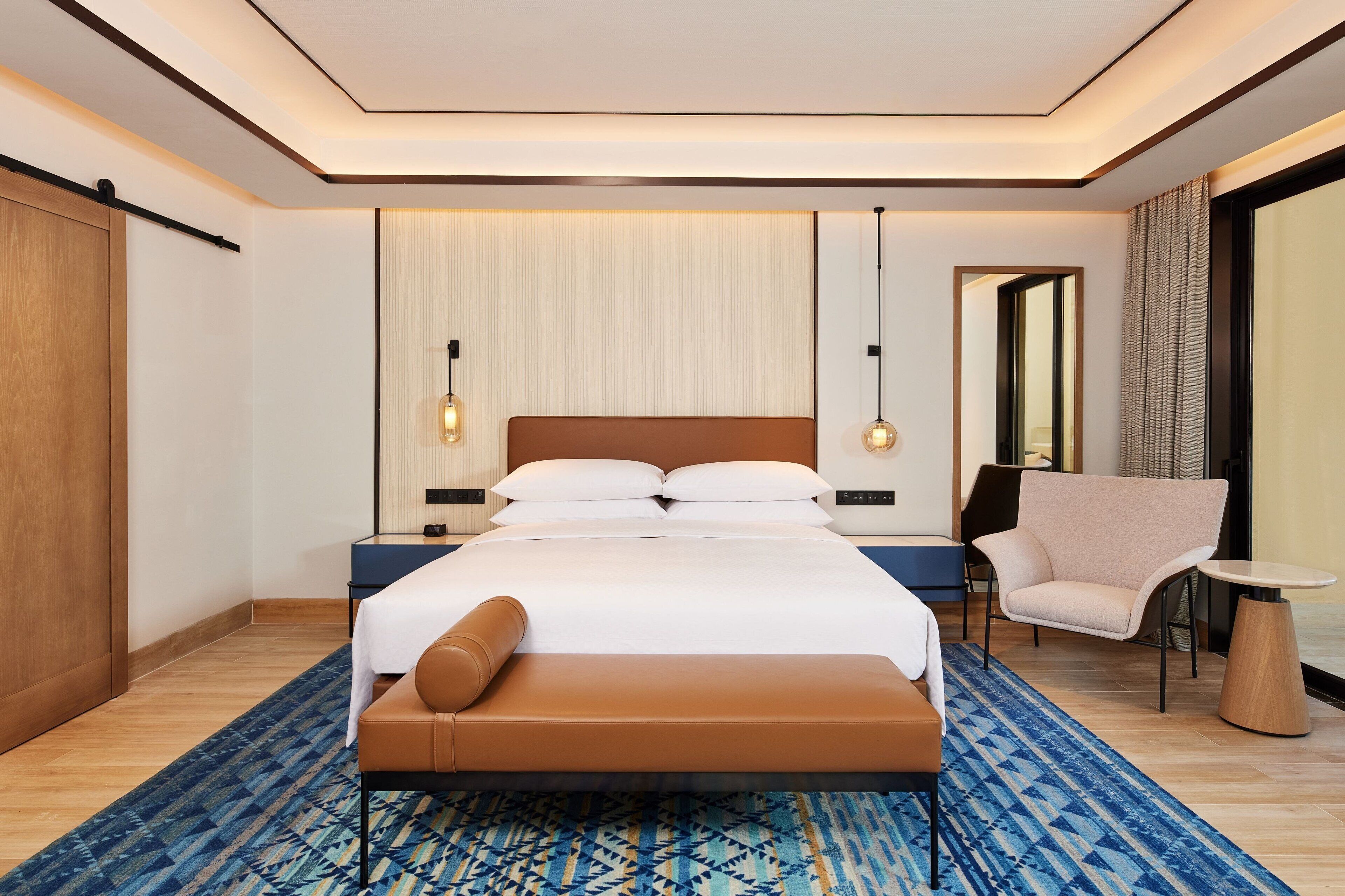 Une chambre d’hôtel moderne dotée d’un grand lit, d’un coin salon avec un fauteuil et une petite table, d’une armoire en bois et d’un grand miroir.