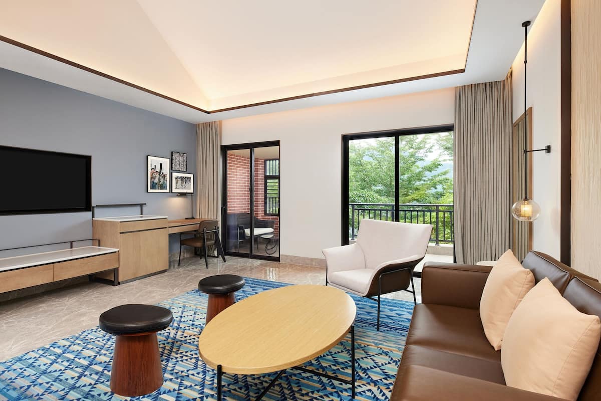 Habitación de hotel moderna con televisión de pantalla plana, sofá, mesa de centro y balcón con vistas a zonas verdes.