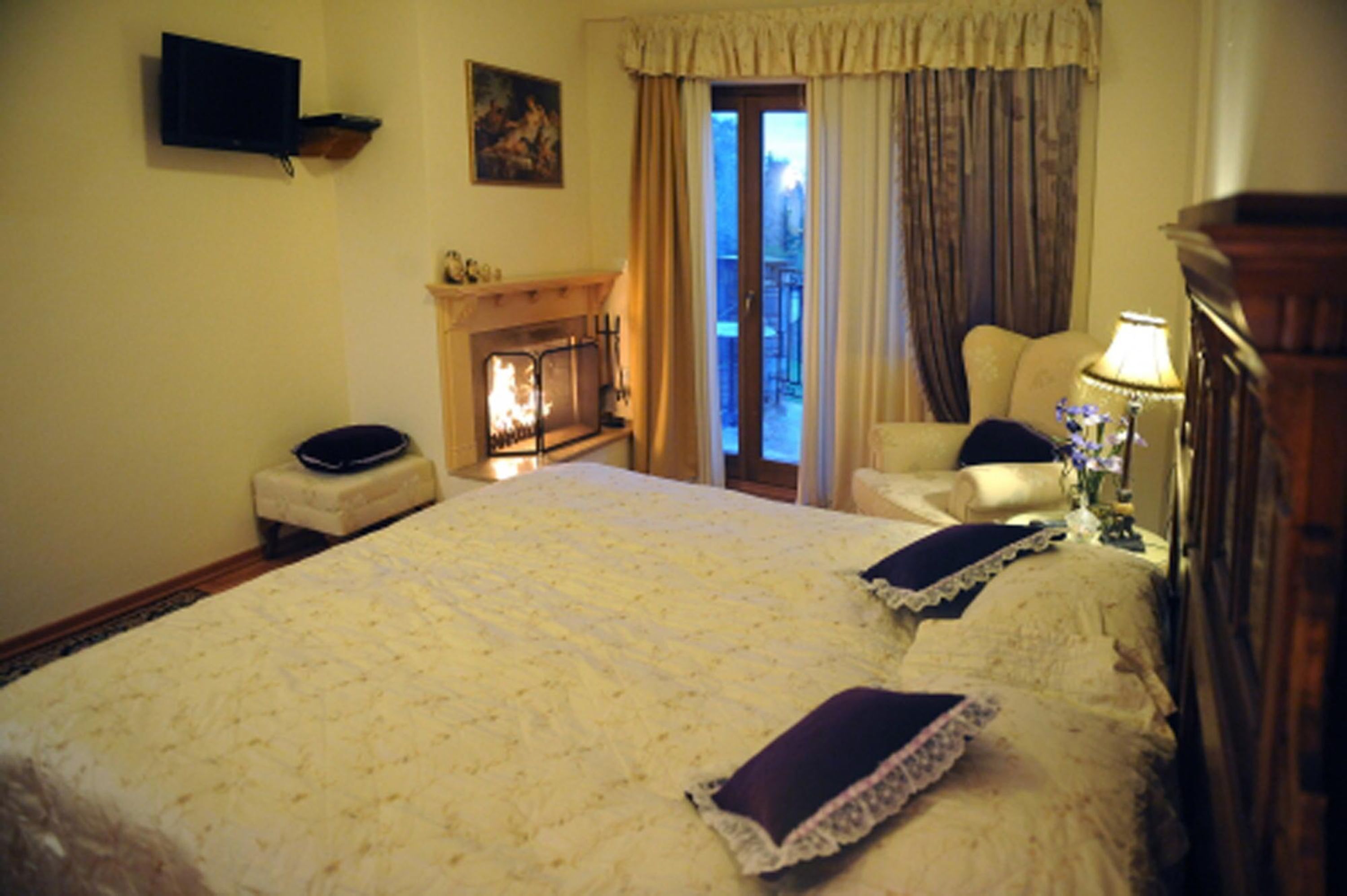 A bedroom with a large bed, a fireplace, a television on the wall, and a balcony with curtains.