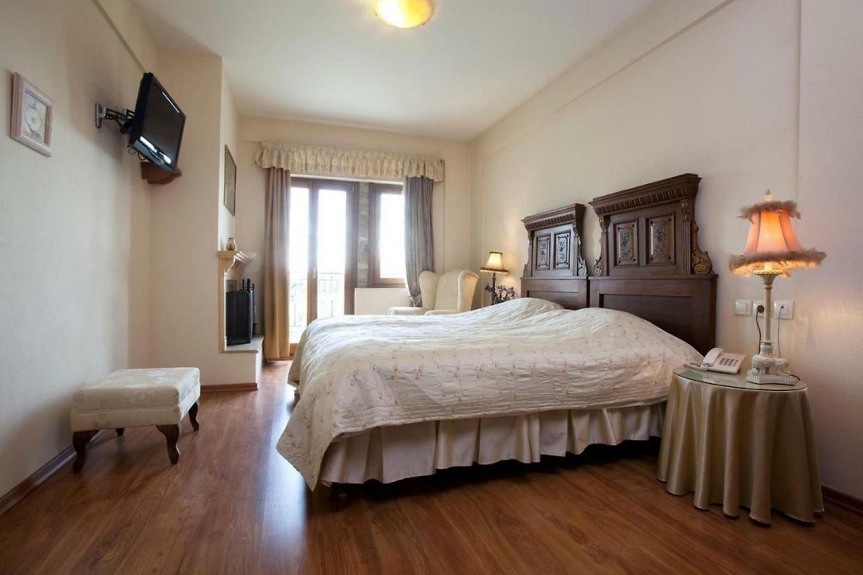 Une chambre avec un grand lit, une table de chevet avec une lampe, un petit tabouret, un téléviseur fixé au mur et une fenêtre avec des rideaux.