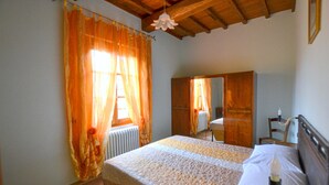 Une chambre avec un plafond en bois, un lit recouvert d’une courtepointe blanche et beige, une table de chevet avec une lampe, et une fenêtre ornée de rideaux orange.