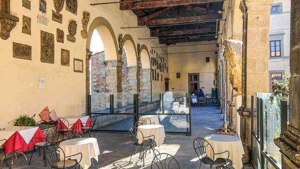 Un café en plein air avec des tables et des chaises, un mur de pierre orné de plaques commémoratives et une vue sur une rue.