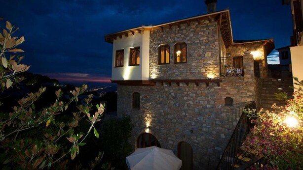 Un edificio de piedra con varias ventanas y un balcón, rodeado de follaje y un atardecer al fondo.