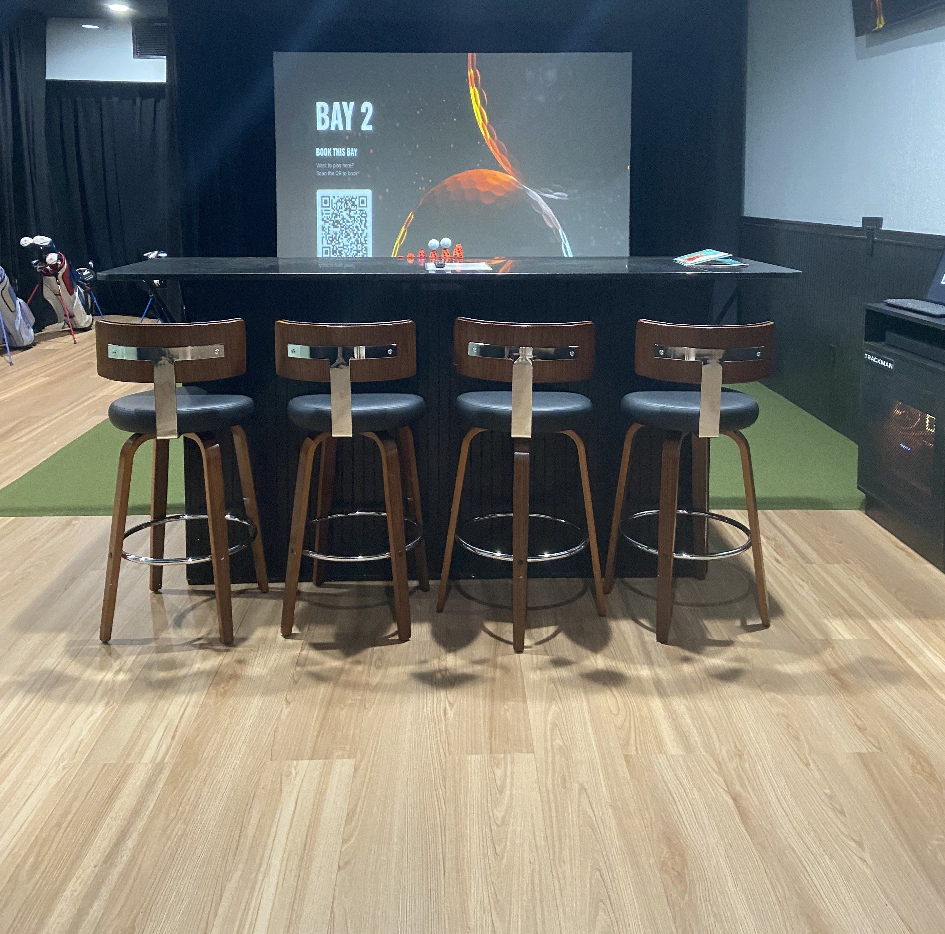 Un espace bar avec un écran affichant la mention « SALLE 2 » et un code QR. On y trouve quatre tabourets de bar en bois avec des pieds en métal.