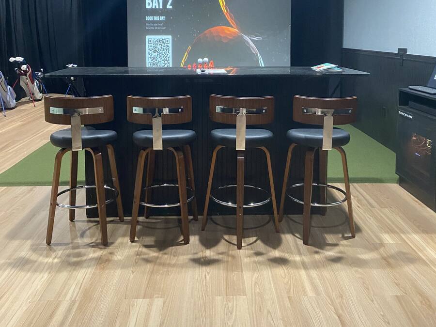 A bar area with a screen displaying "BAY 2" and a QR code. There are four wooden bar stools with metal legs.