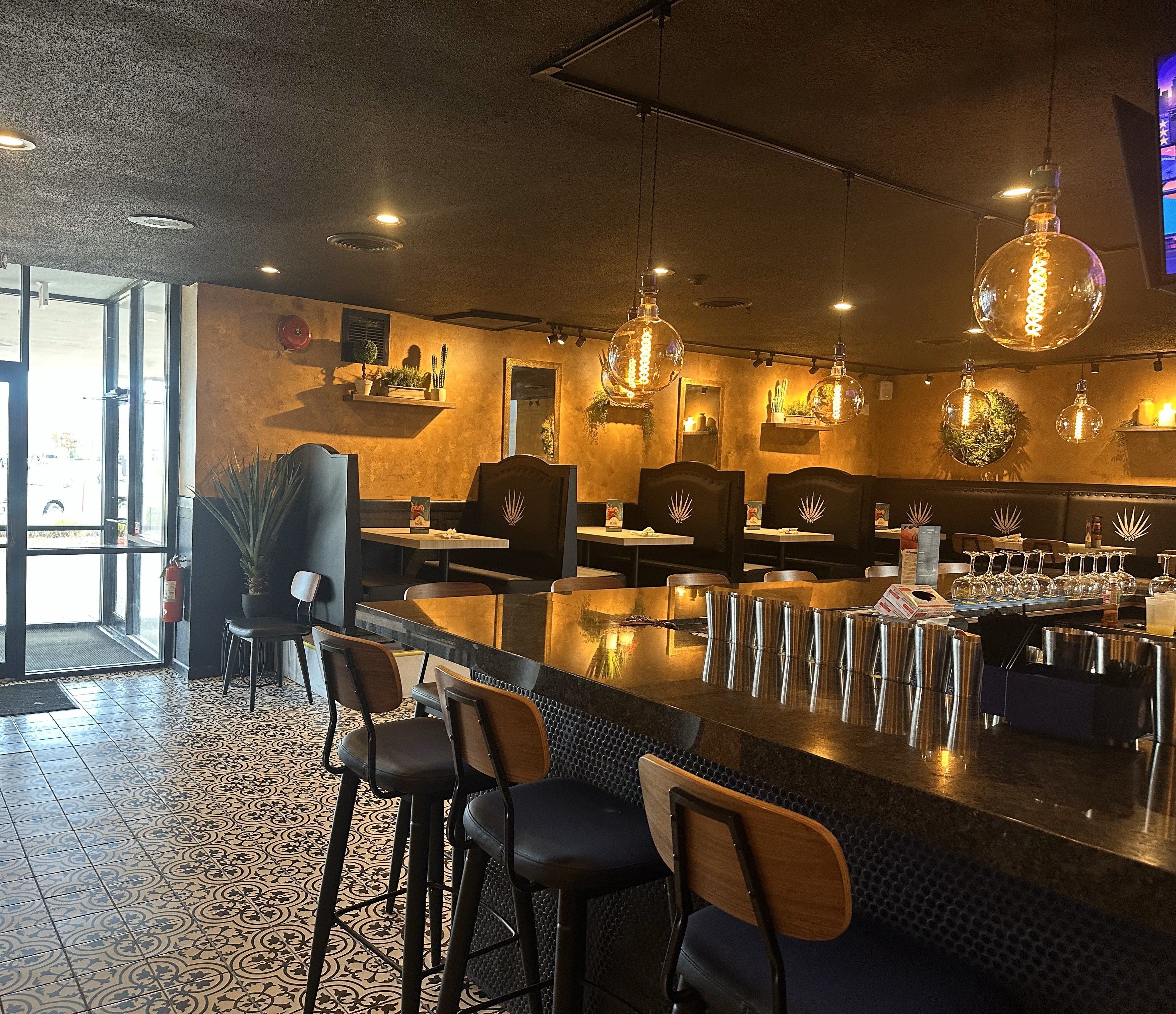 Interior de restaurante con asientos de bar, iluminación colgante y estantes decorativos en la pared.