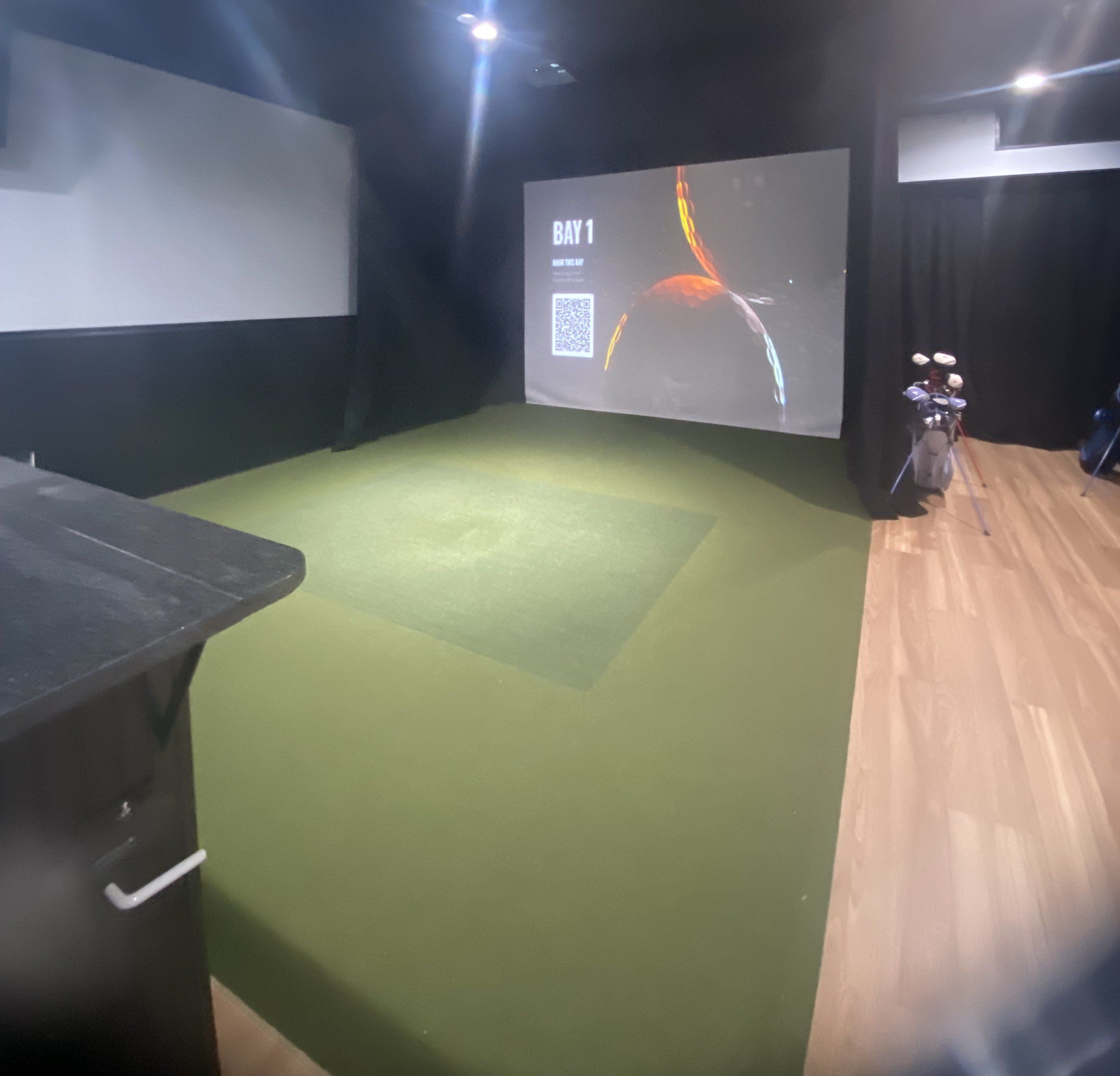 Ein Zimmer mit grünem Teppich, einer Projektionsleinwand, die 'BAY 1' zeigt, und einem Motorrad in der Ecke.