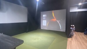A room with a green carpet, a projector screen displaying 'BAY 1', and a motorcycle in the corner.