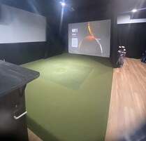 Um quarto com carpete verde, uma tela de projetor exibindo 'BAY 1' e uma motocicleta no canto.