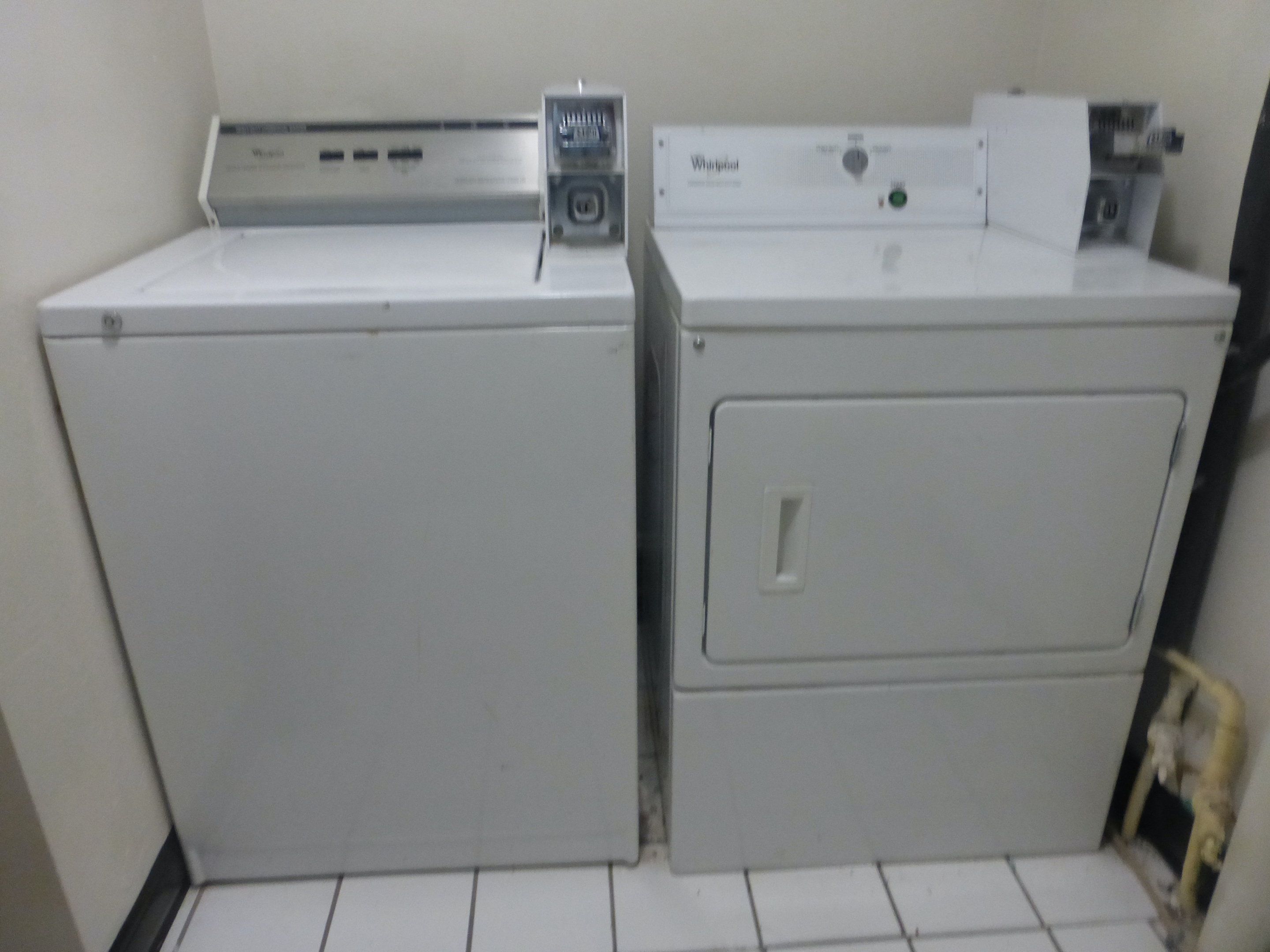 Deux appareils électroménagers blancs, probablement une machine à laver et un sèche-linge, sont placés l’un sur l’autre dans un coin, sur un sol carrelé de carreaux blancs.