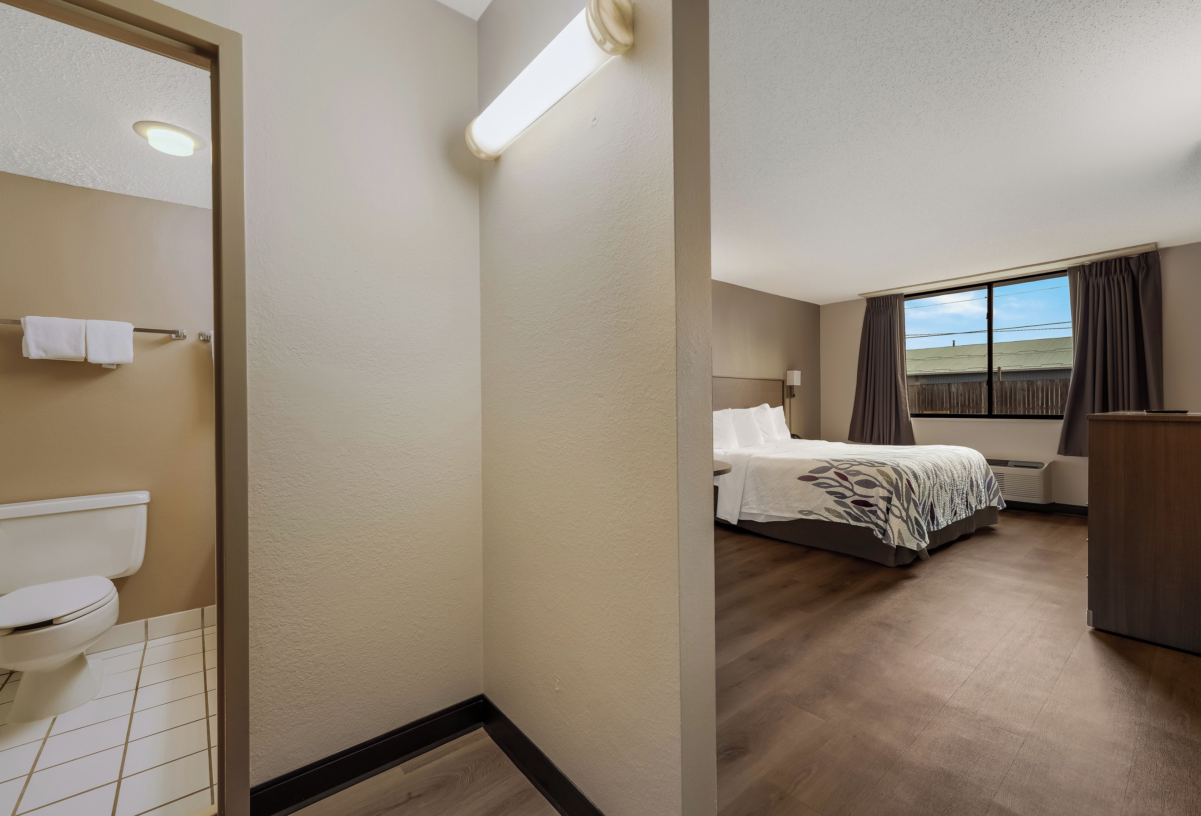 A hotel room with a toilet, a bed with a patterned bedspread, a bedside table, and a window with curtains.