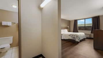 A hotel room with a toilet, a bed with a patterned bedspread, a bedside table, and a window with curtains.