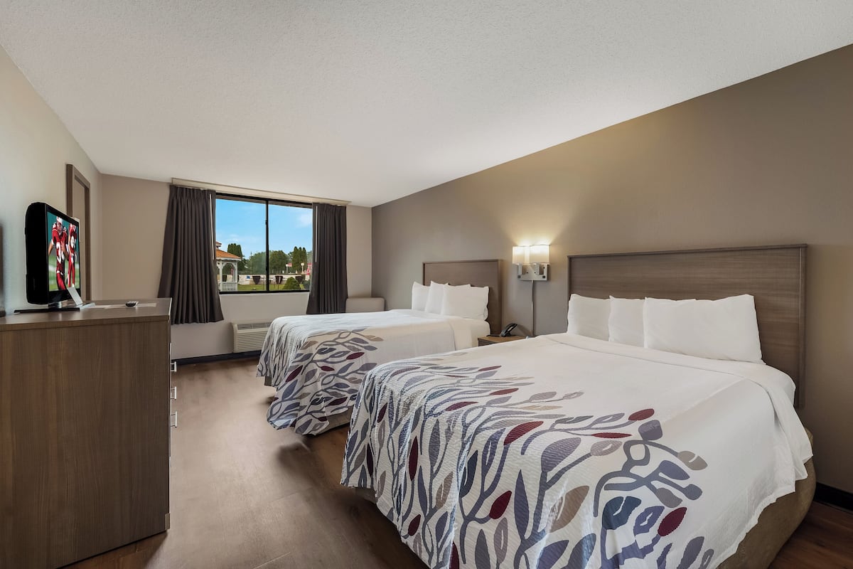 A hotel room with two beds, a desk, a television, and a window with a view of buildings.