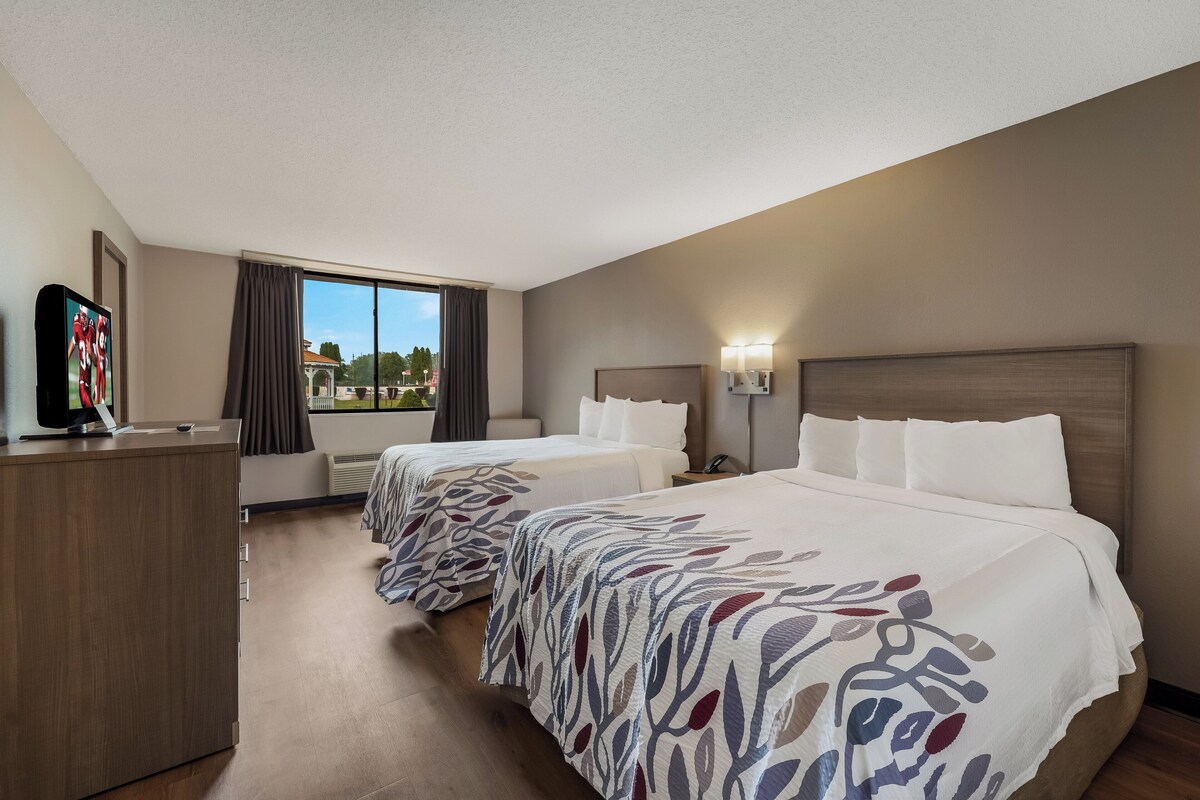 A hotel room with two beds, a desk, a television, and a window with a view of buildings.