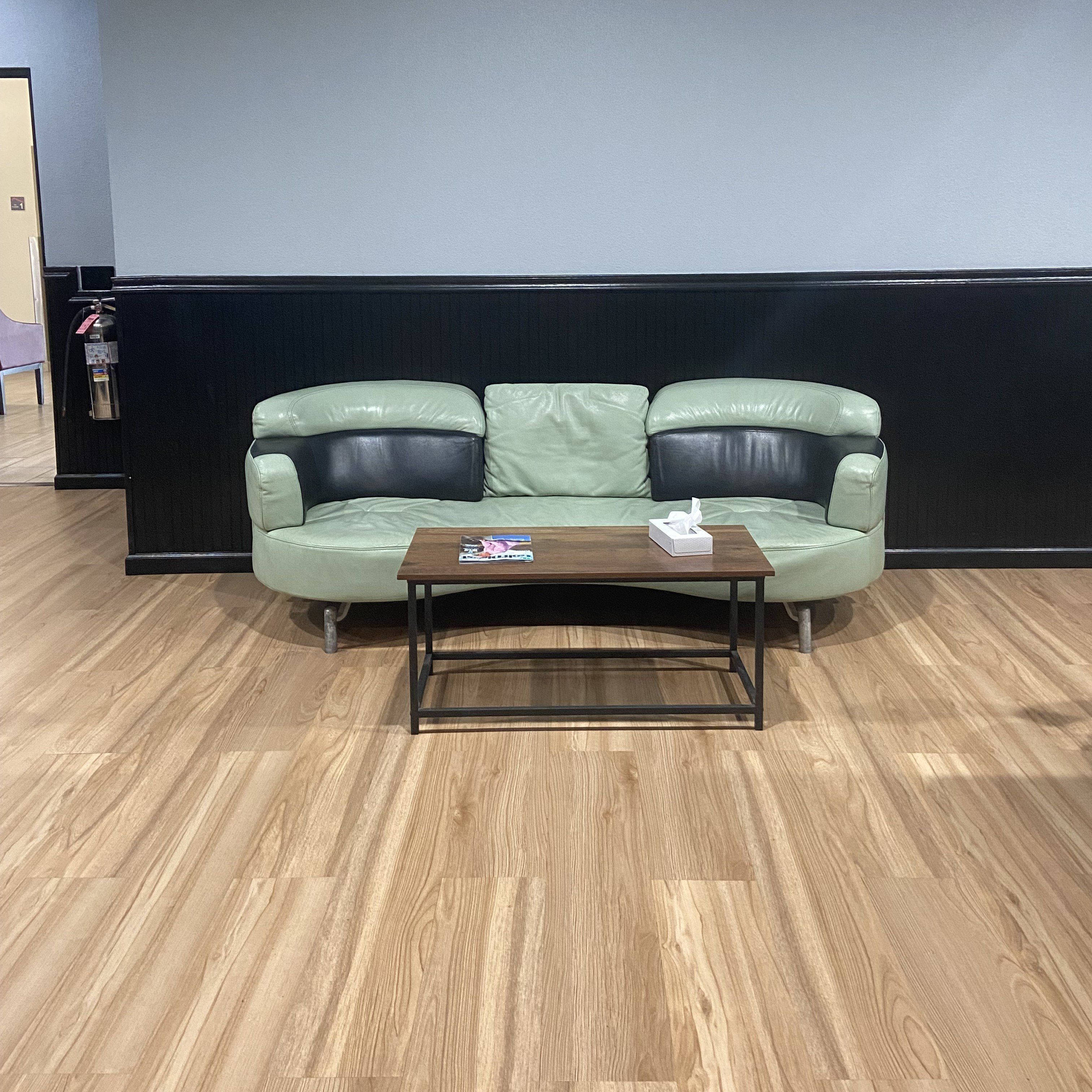 Un canapé en cuir vert, avec une table basse en bois devant, placé contre un mur sombre et sur un plancher en bois.