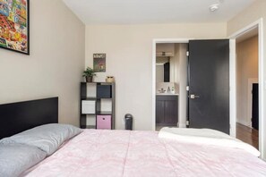Une chambre à coucher avec un lit, une table de chevet, une étagère avec des objets, une porte donnant sur une salle de bain et une affiche accrochée au mur.