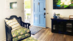 Una stanza con una porta blu, una poltrona nera con rivestimento floreale, un pavimento in legno, un consolle nero con soprammobili e un quadro con fiori sulla parete.