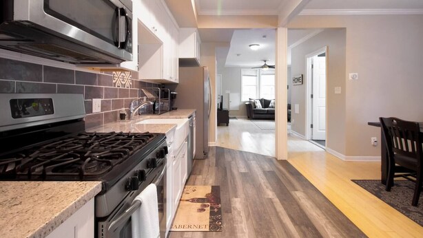 A modern kitchen with a gas stove, microwave, and stainless steel appliances. A wooden floor runs through the space, leading to a living area with a sofa and a coffee table. - Gray Corner Place | Walk Score 90 | Sleeps 9 (Washington)
