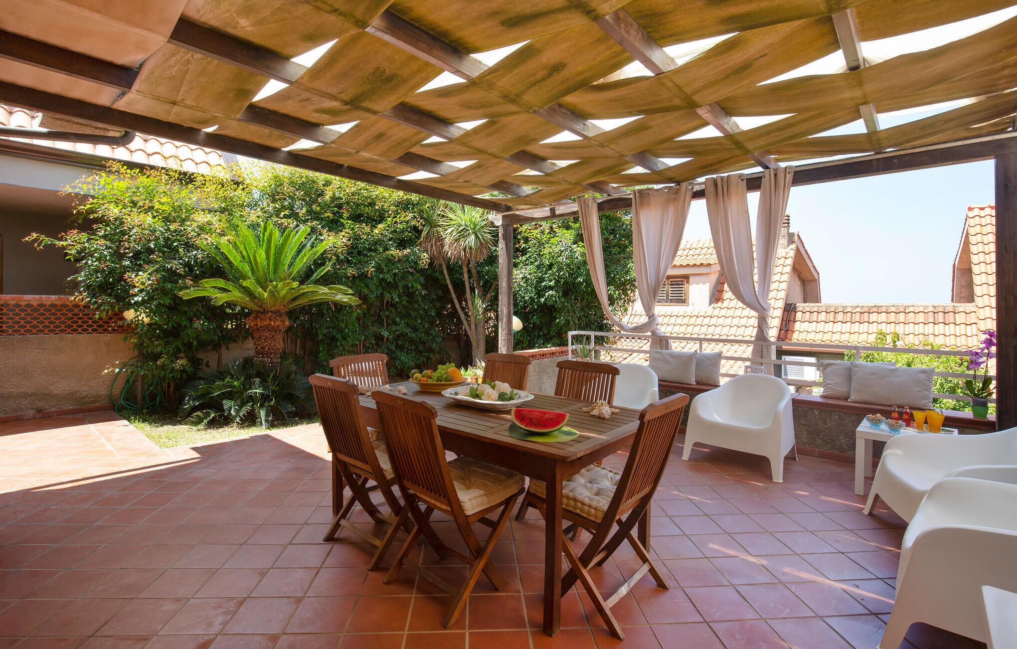 An outdoor patio with a wooden table and chairs, surrounded by greenery and a pergola.