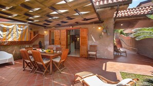 An outdoor patio with wooden furniture, a dining table set with plates and fruits, and a staircase leading to an upper level.