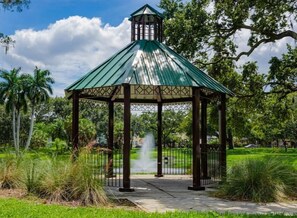 Situated in a serene park-like setting, this gazebo features a decorative lattice design and a green roof, surrounded by lush greenery and a metal fence.