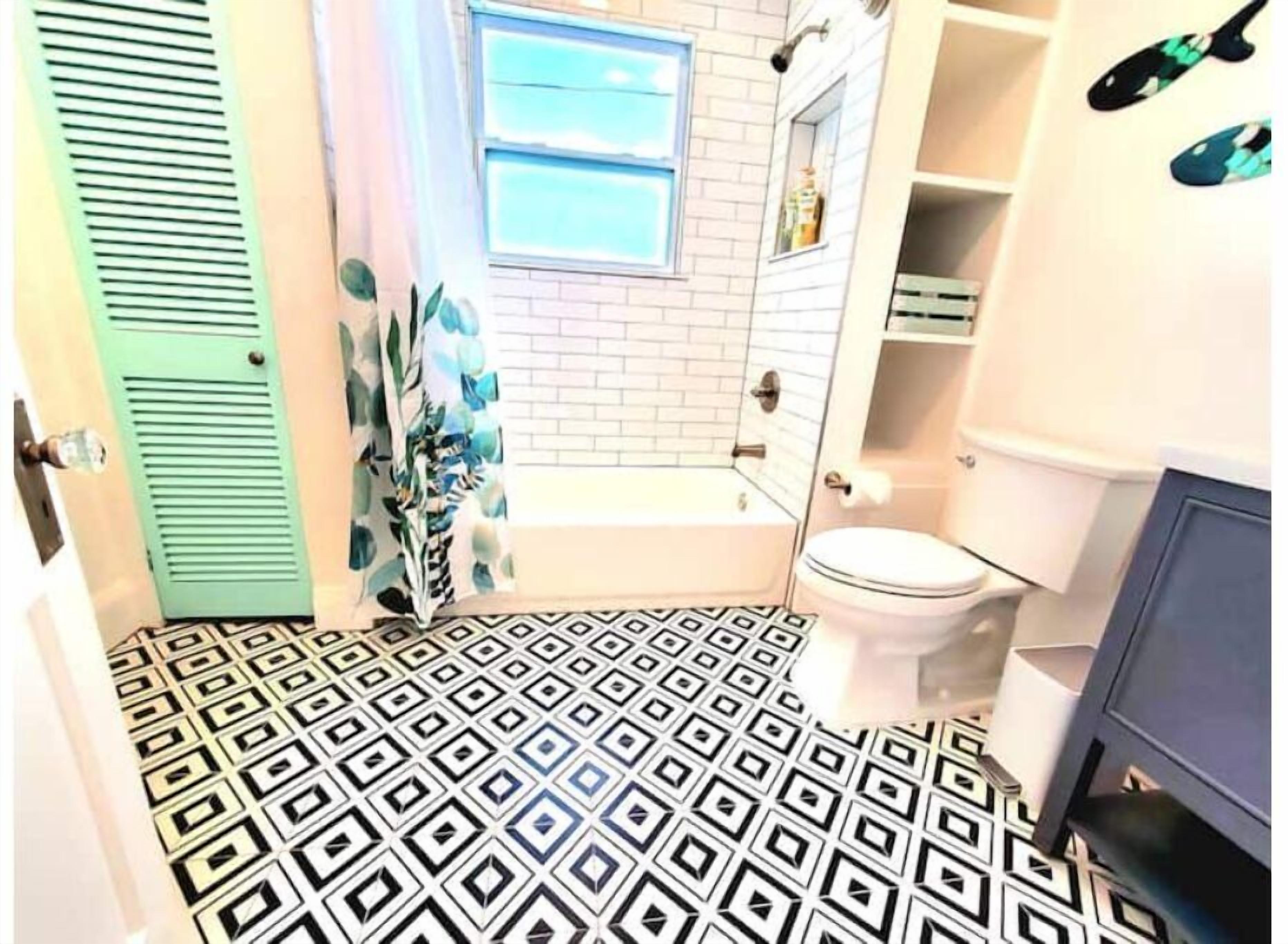 Ein Badezimmer mit einem geometrisch gemusterten Boden, einer weißen Badewanne, einer Toilette und einer grünen Tür.