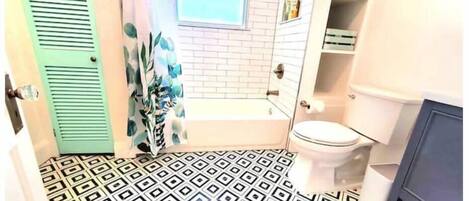 Ein Badezimmer mit einem geometrisch gemusterten Boden, einer weißen Badewanne, einer Toilette und einer grünen Tür.
