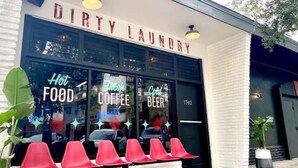 Fachada com uma placa indicando "DIRTY LAUNDRY" e janelas exibindo "COMIDA QUENTE", "CAFÉ FRESCO" e "CERVEJA GELADA".