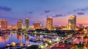 Paisagem urbana ao anoitecer, com arranha-céus, um calhavete com barcos e um calçadão à beira-mar.