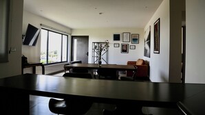 Een moderne eetruimte met een lange tafel, stoelen en een bank. Er hangen afbeeldingen in lijsten aan de muur en een raam zorgt voor natuurlijk licht.