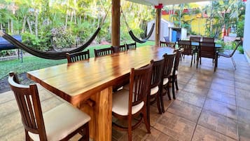 A wooden dining table with chairs, surrounded by a tiled patio with hammocks and a view of a lush garden.