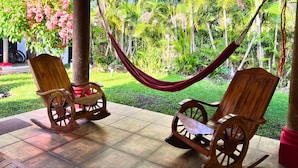 Two wooden rocking chairs with a hammock between them, set on a tiled patio with a lush garden in the background.