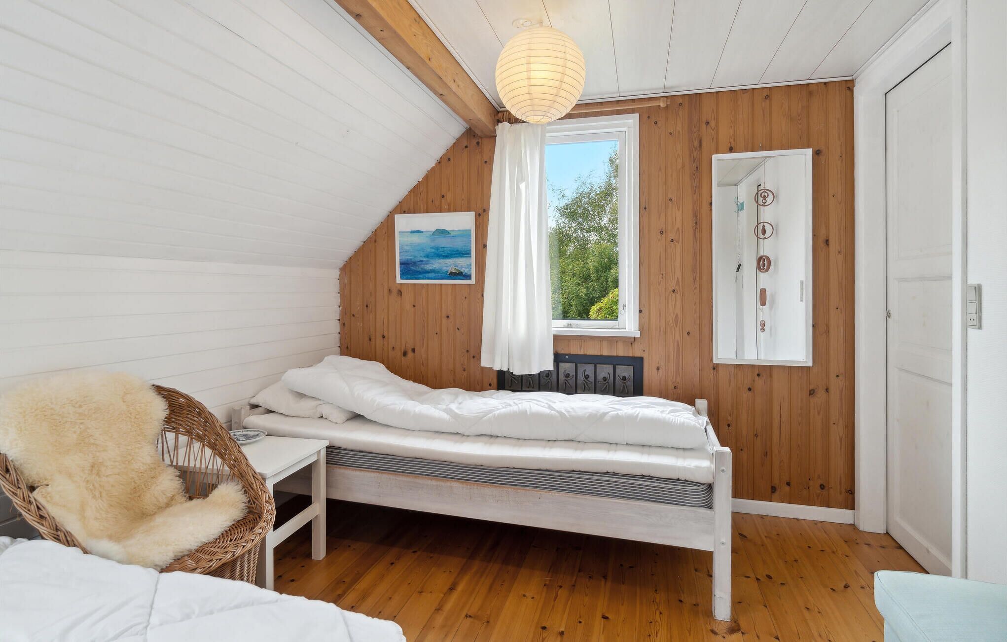 Un dormitorio acogedor con techo inclinado, cama con ropa de cama blanca, silla de mimbre con cojín mullido, una mesita y una ventana con vistas a la vegetación.