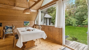 Ein Holzinterieur mit einem für zwei Personen gedecktem Esstisch, einer hängenden Leuchte und Blick auf einen üppigen Garten durch ein großes Fenster.
