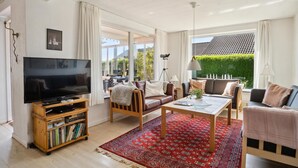 A modern living room with a flat-screen TV, wooden furniture, a red patterned rug, and large windows offering a view of greenery and a tiled roof. - Awesome home in Slagelse with WiFi (Slagelse)