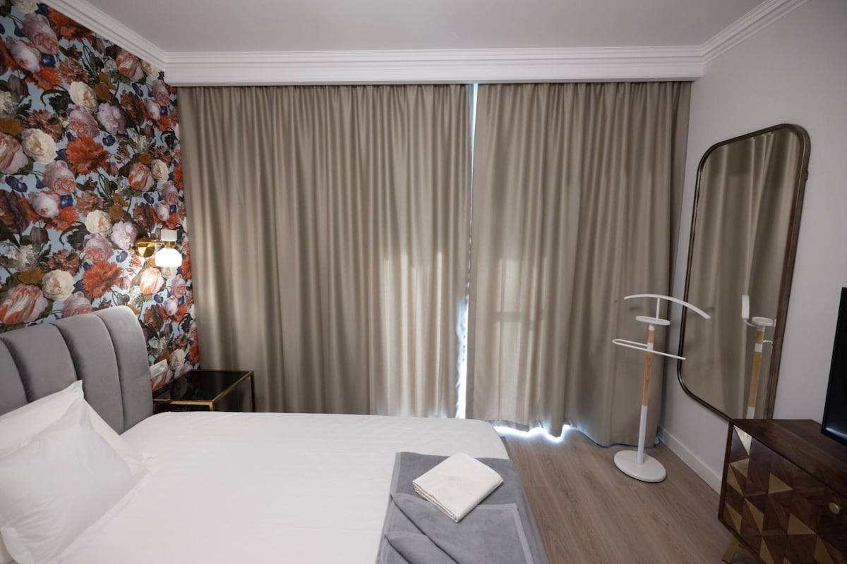 A hotel room with a large bed, a bedside table, a mirror, and a floral wallpaper.