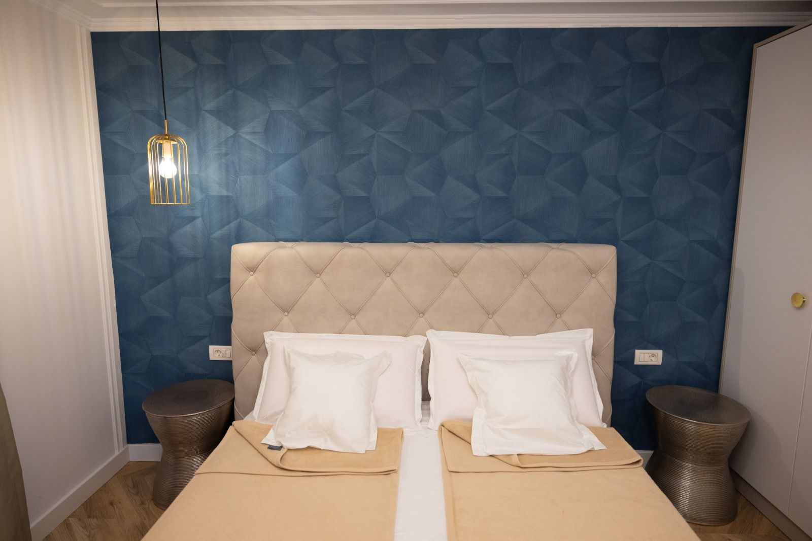 Una camera da letto con una testiera capitonné, due cuscini bianchi, una coperta beige, due comodini e un lampadario.